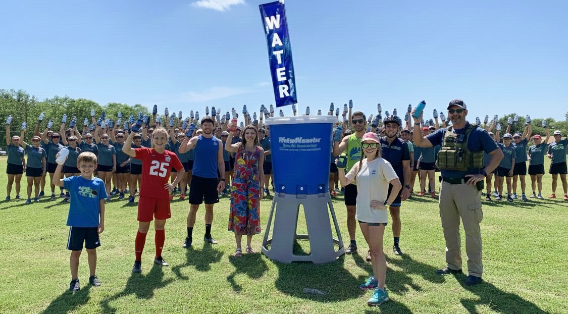 Group of people standing around a water dispenser with 'WATER' sign on a sunny day.
